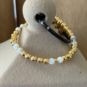 May include: A beaded bracelet featuring a repeating pattern of polished gold-tone metallic spheres and smooth, round white beads. The bracelet is constructed with a flexible cord, showcasing a consistent arrangement of alternating gold and white elements.