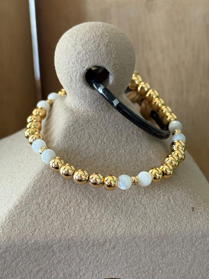 May include: A beaded bracelet featuring a repeating pattern of polished gold-tone metallic spheres and smooth, round white beads. The bracelet is constructed with a flexible cord, showcasing a consistent arrangement of alternating gold and white elements.