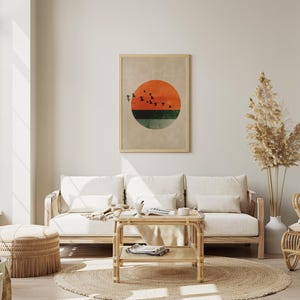 May include: A living room scene featuring a framed abstract art print with a sunset design in orange, green, and black. The room includes a light wood sofa with white cushions, a woven coffee table, and a round jute rug. Decorative pampas grass adds a natural touch.