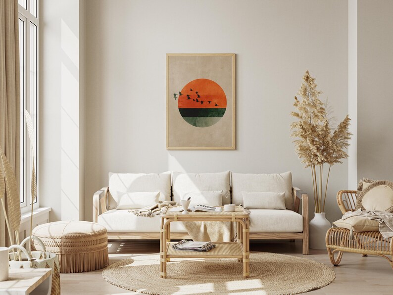 May include: A living room scene featuring a framed abstract art print with a sunset design in orange, green, and black. The room includes a light wood sofa with white cushions, a woven coffee table, and a round jute rug. Decorative pampas grass adds a natural touch.