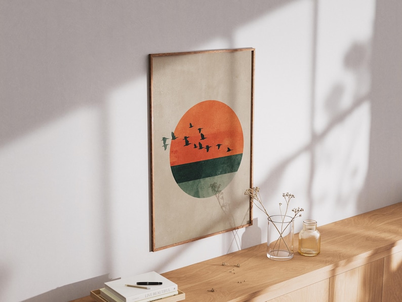 May include: A framed art print featuring a minimalist design of a sunset with birds in flight. The artwork showcases an orange sun, a green and blue horizon, and a flock of birds against a neutral background. The frame is a warm brown.