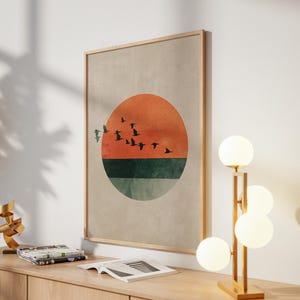 May include: A framed wall art print featuring a sunset design with a vibrant orange and green color scheme. Silhouetted birds fly across the orange sun. The artwork is displayed on a light-colored wall, next to a modern lamp.