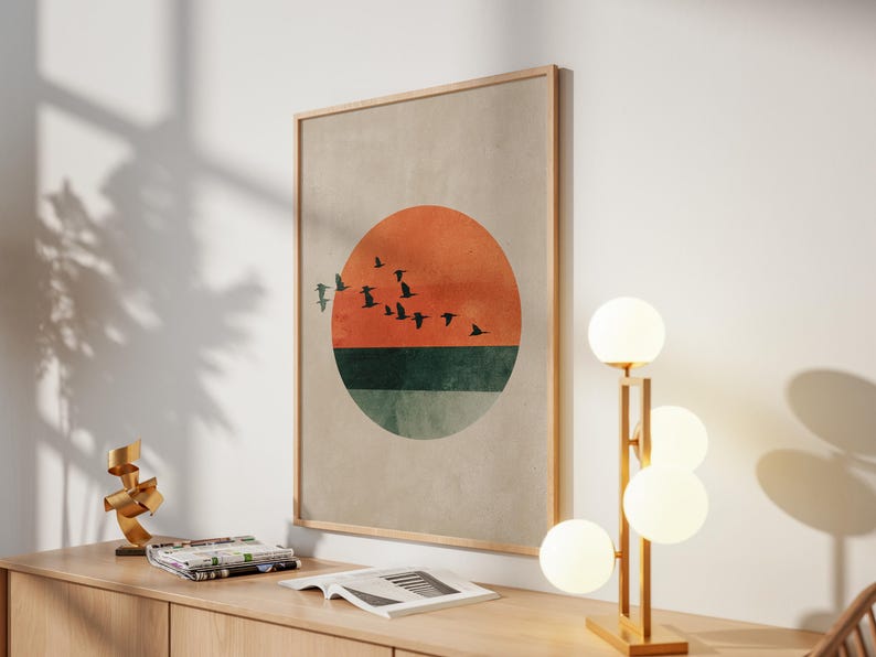 May include: A framed wall art print featuring a sunset design with a vibrant orange and green color scheme. Silhouetted birds fly across the orange sun. The artwork is displayed on a light-colored wall, next to a modern lamp.
