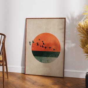May include: A framed art print featuring a minimalist design. The artwork showcases a large orange circle representing the sun, with a flock of birds in silhouette flying across it. Below the sun is a green and teal section. The background is a neutral beige.