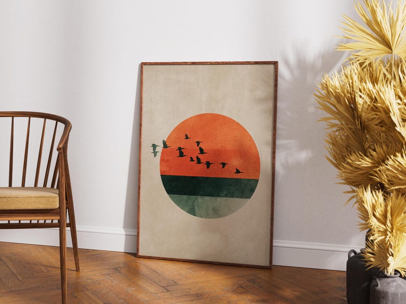 May include: A framed art print featuring a minimalist design. The artwork showcases a large orange circle representing the sun, with a flock of birds in silhouette flying across it. Below the sun is a green and teal section. The background is a neutral beige.