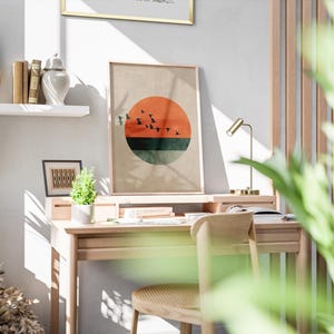 May include: A wooden desk with a framed print featuring a sunset design. The print has a red circle, a green stripe, and black birds. A small desk lamp, books, and a small plant are also visible.