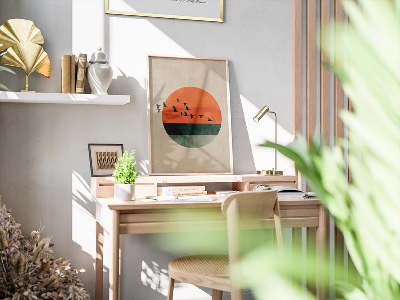 May include: A wooden desk with a framed print featuring a sunset design. The print has a red circle, a green stripe, and black birds. A small desk lamp, books, and a small plant are also visible.