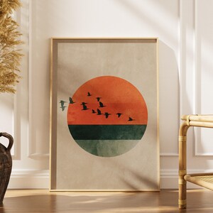 May include: Framed art print featuring a minimalist sunset design. The artwork showcases a vibrant orange sun, a green horizon, and silhouetted birds in flight against a neutral background. The print is encased in a light-colored wooden frame.