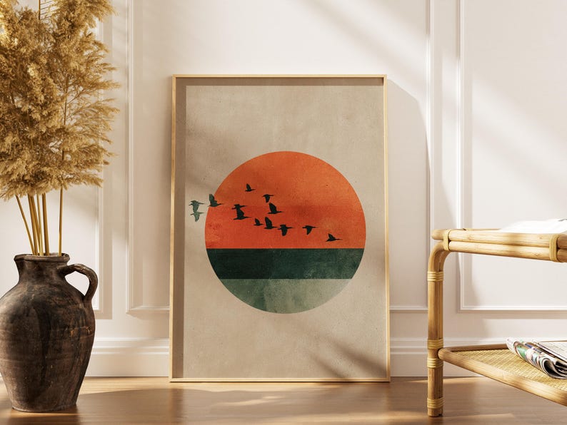 May include: Framed art print featuring a minimalist sunset design. The artwork showcases a vibrant orange sun, a green horizon, and silhouetted birds in flight against a neutral background. The print is encased in a light-colored wooden frame.
