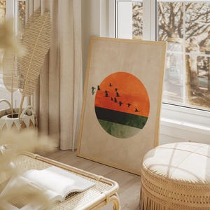 May include: A framed art print featuring a minimalist design of a sunset with birds in flight. The artwork has a large orange and green circle with a flock of black birds. The frame is light brown, and the print is leaning against a wall.