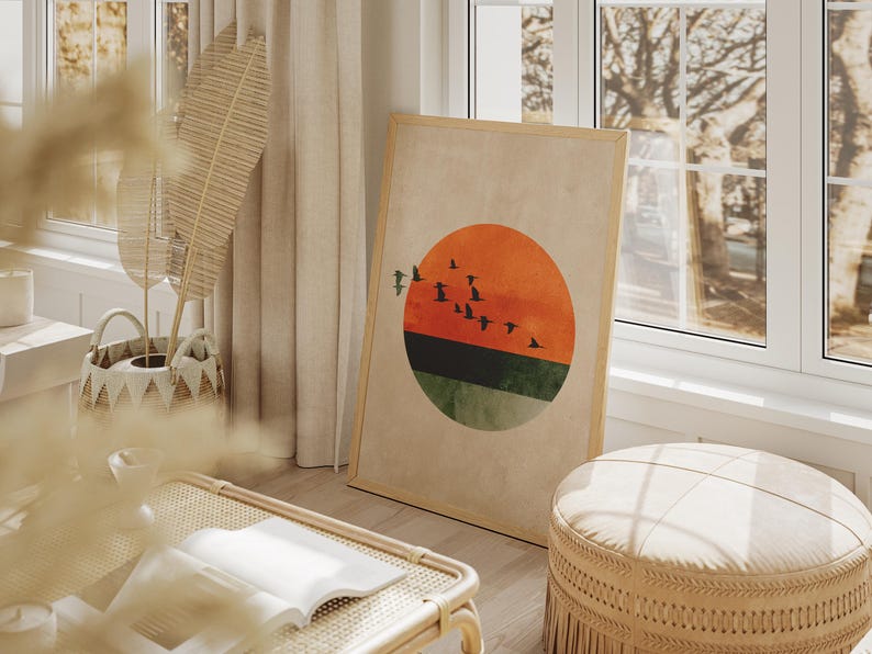 May include: A framed art print featuring a minimalist design of a sunset with birds in flight. The artwork has a large orange and green circle with a flock of black birds. The frame is light brown, and the print is leaning against a wall.