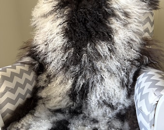 May include: A long-haired sheepskin rug featuring a mottled pattern of black, charcoal, and white curls. The thick, textured fleece is draped over a chair upholstered in a grey and white chevron patterned fabric, showcasing natural variations.