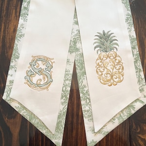May include: Two white decorative table runners with green and white patterned borders. One runner features an embroidered gold and teal letter "S", the other a gold and green pineapple design. The runners are on a dark wood surface.