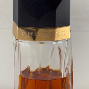 May include: A vintage perfume bottle with a black geometric cap and a gold band that reads "KNOWING". The clear glass bottle contains a deep amber liquid. The bottle has a unique, faceted design.