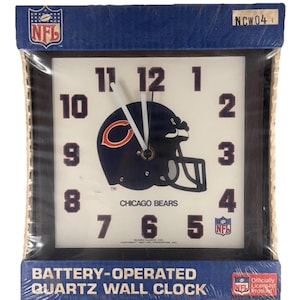 Könnte beinhalten: Quadratische Wanduhr mit dem Logo der Chicago Bears, einem marineblauen Footballhelm mit einem orangefarbenen C. Die Uhr hat schwarze Zeiger, schwarze Ziffern und das NFL-Logo. Die Uhr befindet sich in der Originalverpackung.