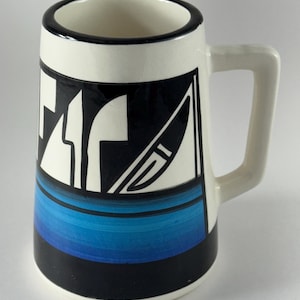 May include: A ceramic mug with a cream-colored base and a black rim. The exterior features a graphic design with black and white abstract shapes above a horizontal band of gradient blue stripes, finished with a solid black base.