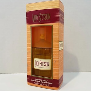 May include: A rectangular box of Lady Stetson cologne. The box is orange and has a burgundy stripe with the brand name. The cologne bottle is visible through a window. The text "COLOGNE SPRAY" and "1.5 FL OZ 44 mL" are printed on the box.