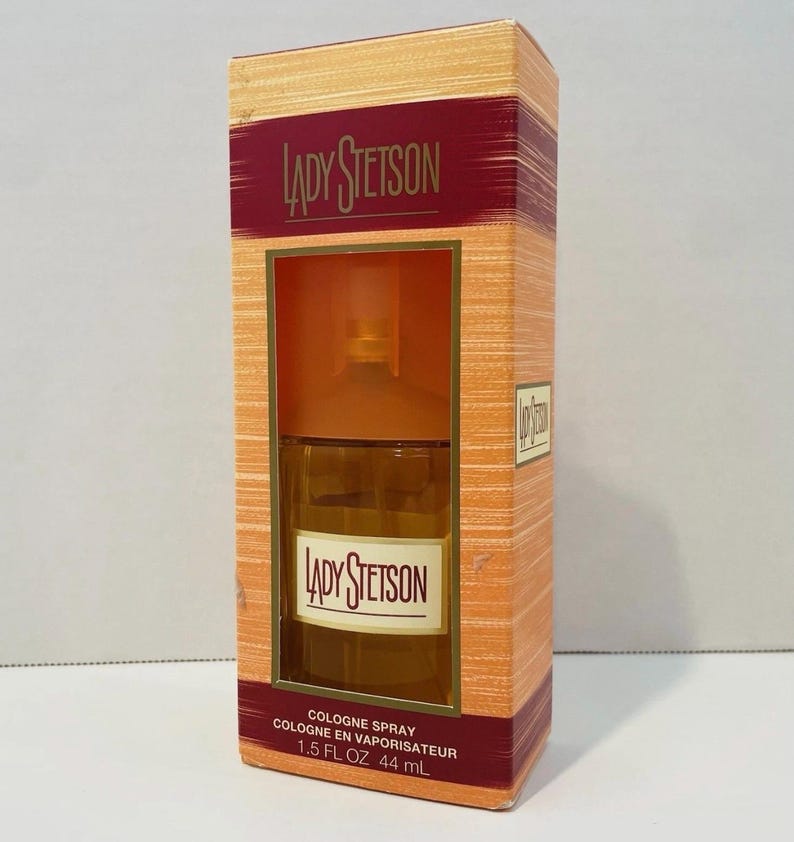 May include: A rectangular box of Lady Stetson cologne. The box is orange and has a burgundy stripe with the brand name. The cologne bottle is visible through a window. The text "COLOGNE SPRAY" and "1.5 FL OZ 44 mL" are printed on the box.