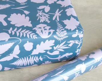 Peut inclure: Un rouleau de papier cadeau et une boîte emballée présentant un fond bleu sarcelle foncé avec un motif répétitif de silhouettes lavande clair représentant divers éléments forestiers dont des champignons, fougères, feuilles de chêne, libellule et lézard.