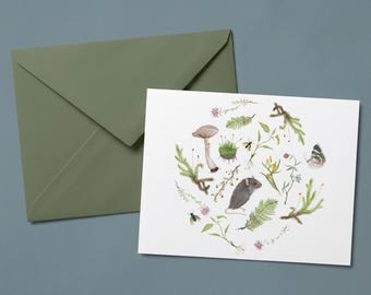 Peut inclure: Une carte de vœux blanche présentant une illustration circulaire à l'aquarelle d'une souris grise entourée d'éléments botaniques variés, dont un champignon, des fougères vertes, des petites fleurs jaunes, de la mousse, un papillon et deux insectes, avec une enveloppe vert sauge.