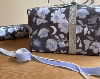 Peut inclure: Une boîte cadeau rectangulaire enveloppée dans du papier gris foncé avec un motif de fleurs bleu clair et de croissants de lune blancs, nouée avec un ruban argenté scintillant, accompagnée d'un ruban de velours violet clair.