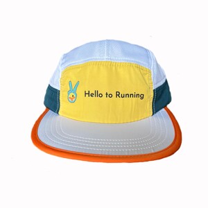 Puede incluir: Gorra de running con parte superior de malla blanca, panel frontal amarillo con el texto "Hello to Running" y un gráfico de signo de la paz. La visera es blanca con ribete naranja y los laterales son turquesa.
