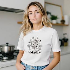 May include: White t-shirt featuring a floral and butterfly design with the text "5 Solas". The graphic includes the phrases "Sola Scriptura", "Sola Fide", "Solus Christus", and "Sola Gratia".