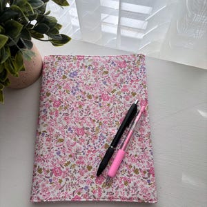 May include: A rectangular fabric notebook cover featuring a dense floral pattern with pink, purple, and green blossoms on a white background. A black ballpoint pen and a pink gel pen are placed diagonally across the front cover.