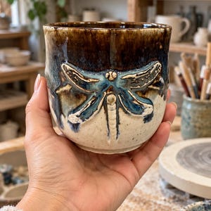 May include: A ceramic mug featuring a raised, sculpted dragonfly design on the front. The upper portion of the mug has a dark brown and blue drip glaze, while the lower portion is a speckled cream color. The dragonfly is detailed in blue and white.
