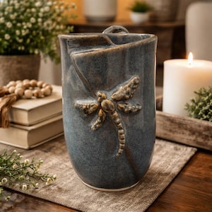 May include: A blue ceramic vase with a dragonfly design. The vase has a textured surface and a handle. The dragonfly is in shades of brown and tan. The vase is sitting on a brown surface.