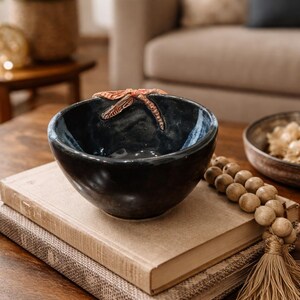 May include: A dark blue ceramic bowl with a dragonfly accent, resting on two stacked books. The bowl has a glossy finish and a decorative dragonfly on the rim. A wooden beaded strand and a second bowl are also visible.