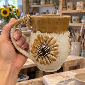 May include: A ceramic mug with a speckled cream-colored body and a brown glazed rim. The mug features a large, three-dimensional sunflower relief on the side and a decorative, knotted handle. The surface has a glossy finish throughout.