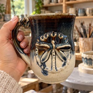 May include: A ceramic mug featuring a raised, sculpted dragonfly emblem on the front. The mug has a two-tone glaze, with a dark blue-black finish on the upper portion transitioning into a speckled cream color on the lower body and handle.