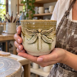May include: A ceramic mug featuring a raised, sculpted dragonfly design on the front. The mug has a speckled off-white base glaze with a dripping, moss-green and earthy brown glaze accent around the rim and over the dragonfly relief.