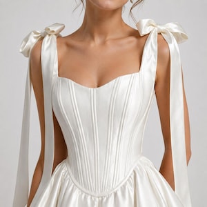 White satin corset dress for women featuring structured boning, square neckline, bow straps, and voluminous short skirt.