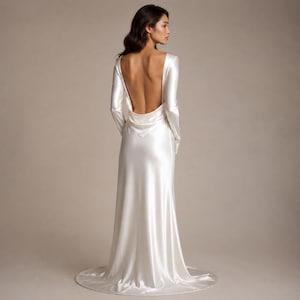 May include: A floor-length, long-sleeved gown in a lustrous white satin fabric. The garment features a deep, open back design with a draped cowl neckline detail at the lower back, long fitted sleeves, and a flowing skirt that pools slightly at the floor.