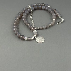 May include: A grey beaded necklace with a silver pendant. The necklace features small, round, grey beads strung together with a silver chain and clasp. The pendant is a hammered silver disc.