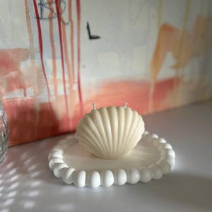 Könnte beinhalten: Eine cremefarbene, muschelförmige Kerze mit zwei Dochten steht auf einem weißen Teller mit Perlenrand. Der Hintergrund zeigt ein abstraktes Gemälde mit roten und orangen Farbtönen.
