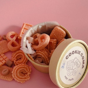 May include: A beige container with a lid, filled with orange wax melts shaped like biscuits and pretzels. The container is open, revealing the contents. The lid has the words "COOKIE COMA" and "wax melts" printed on it. The background is pink.