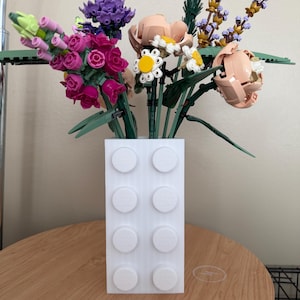 Pode incluir: Um vaso branco de blocos Lego cheio de flores Lego coloridas. As flores incluem rosas cor-de-rosa, lavanda roxa e margaridas brancas com centros amarelos. O vaso está numa mesa de madeira.