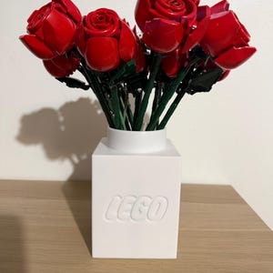 Pode incluir: Um buquê de rosas vermelhas de plástico, dispostas em um vaso branco quadrado. O vaso tem a palavra "LEGO" em relevo na frente. As rosas têm caules verdes e estão em plena floração.