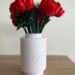 Pode incluir: Um vaso LEGO branco contém um buquê de rosas vermelhas LEGO com caules e folhas verdes. O vaso tem o logotipo LEGO. O arranjo está em uma superfície de madeira marrom clara.