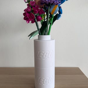 Pode incluir: Um vaso LEGO branco com o logotipo LEGO em relevo duas vezes, segurando um buquê de flores LEGO coloridas. As flores incluem rosas cor-de-rosa, flores roxas e azuis e caules verdes, criando uma exibição vibrante.