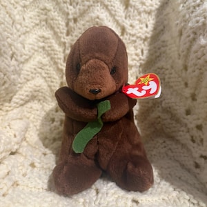 May include: A brown plush otter toy with a green ribbon and a red tag. The stuffed animal has a small black nose and is sitting upright. The tag is red with a white star and the word "BEANIE" on it.