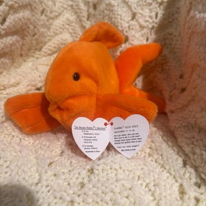 May include: An orange plush Goldie the fish Beanie Baby toy. The toy has a heart-shaped tag attached. The plush fish is on a cream-colored, textured surface. The tag reads "Goldie" and includes a poem.