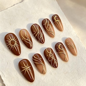 May include: A set of ten almond-shaped press-on nails featuring a brown and beige color palette. The designs include gold raised sun, star, and crescent moon motifs, intricate swirling gold lines, and small embedded clear rhinestones.