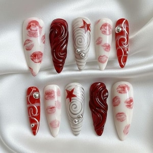 以下が含まれることがあります： 10本の細長いアーモンド型の人工爪のセット。デザインには、白地に赤い唇のプリント、渦巻く赤い模様、赤い星とシルバーの渦巻き、シルバーの装飾が含まれます。白いサテンの表面に配置されています。