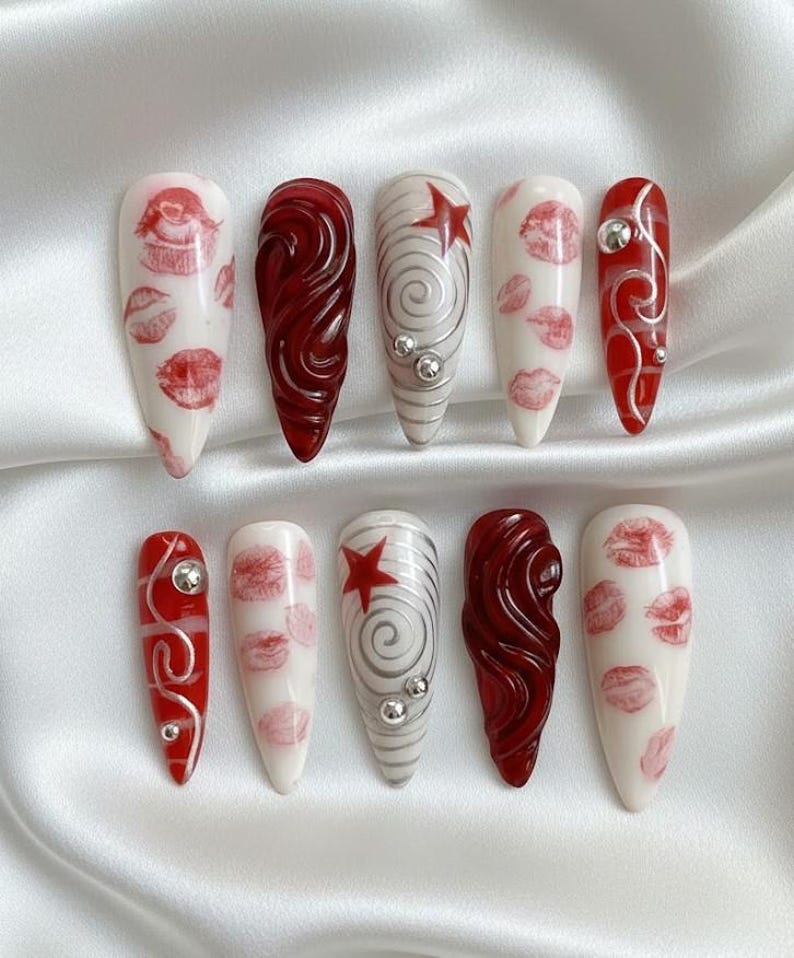 以下が含まれることがあります： 10本の細長いアーモンド型の人工爪のセット。デザインには、白地に赤い唇のプリント、渦巻く赤い模様、赤い星とシルバーの渦巻き、シルバーの装飾が含まれます。白いサテンの表面に配置されています。