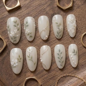May include: A set of ten almond-shaped press-on nails in a creamy off-white finish. Each nail features delicate 3D floral accents, painted green leaves, thin gold metallic lines, and small white pearl embellishments arranged in various patterns.