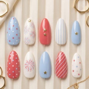 May include: A collection of ten artificial nails in various designs. The nails feature red, white, and blue colors with patterns like polka dots, stripes, fireworks, and star accents. The nails are oval-shaped and arranged on a light-colored surface.